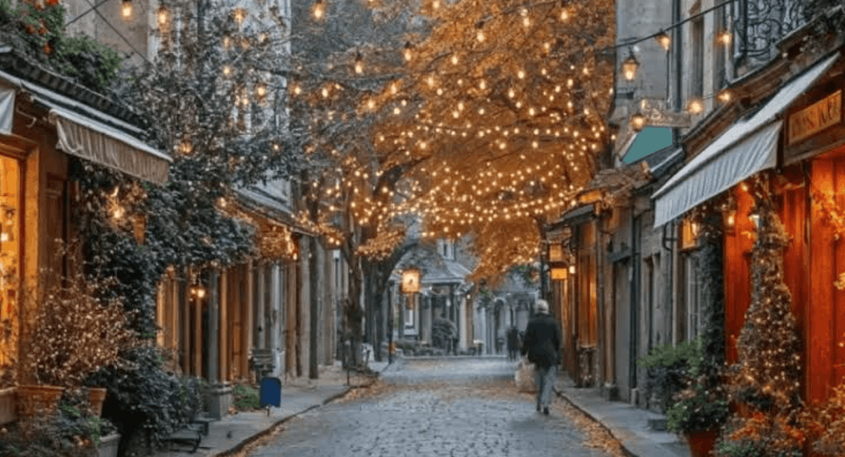 A cobblestone street lined with storefronts and trees decorated with warm string lights in autumn.