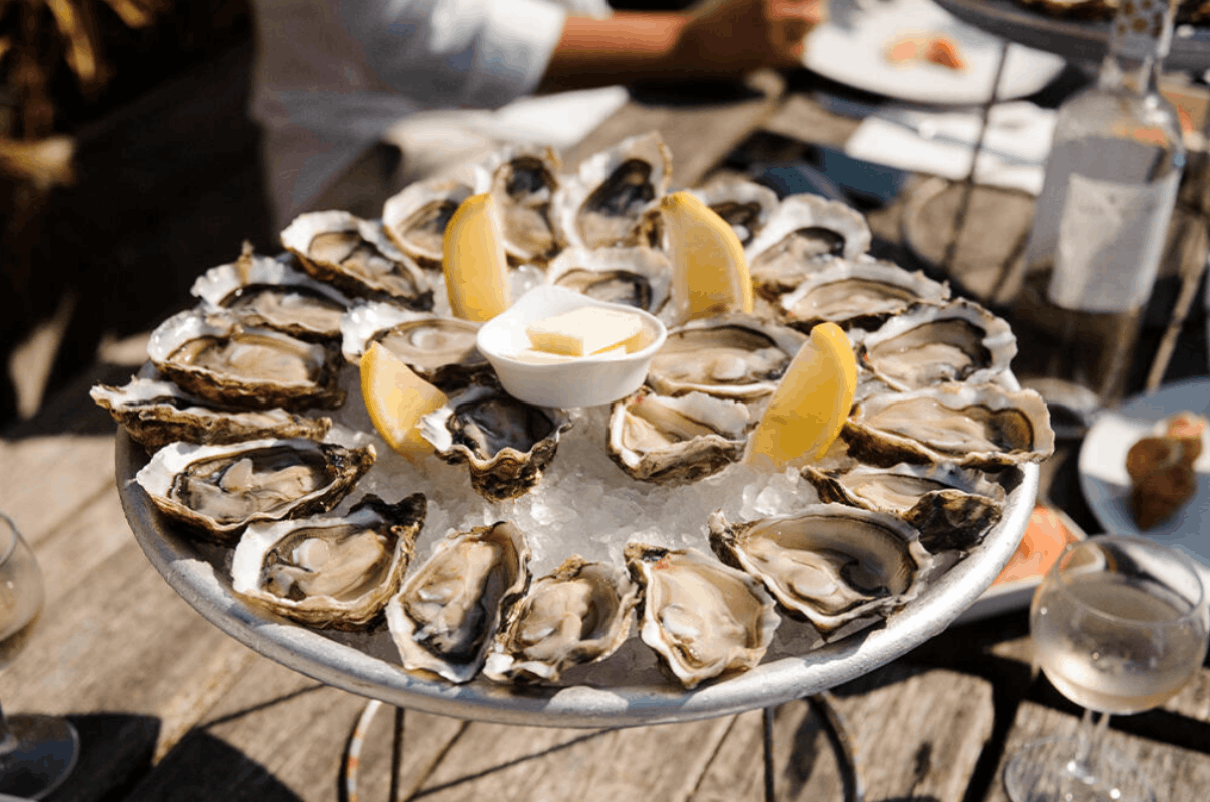 A platter of fresh raw oysters on ice with lemon wedges and butter served at an outdoor table 