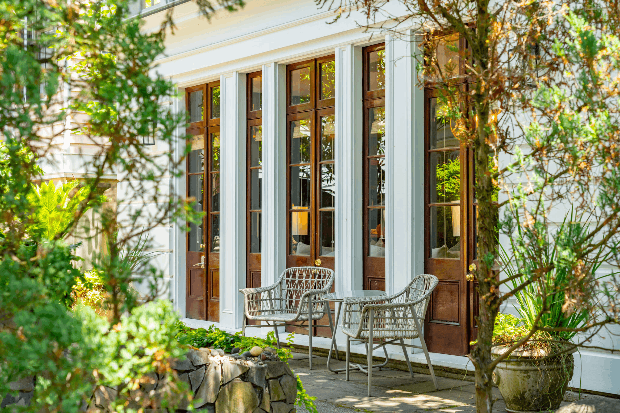 A cozy outdoor patio featuring two woven wicker chairs and a small round table in front of large glass-paneled wooden doors, surrounded by lush green trees and plants.