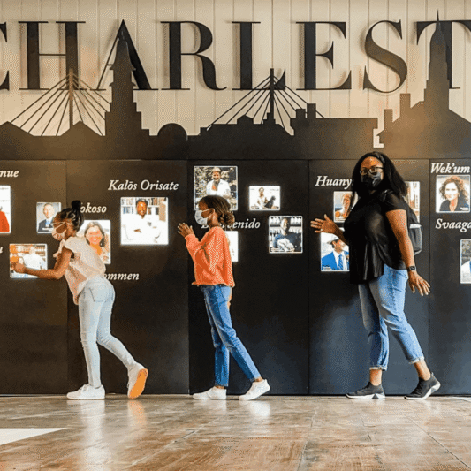A family of four walking in a line in front of a "Welcome to Charleston" display with a city skyline silhouette and multi-language greetings.
