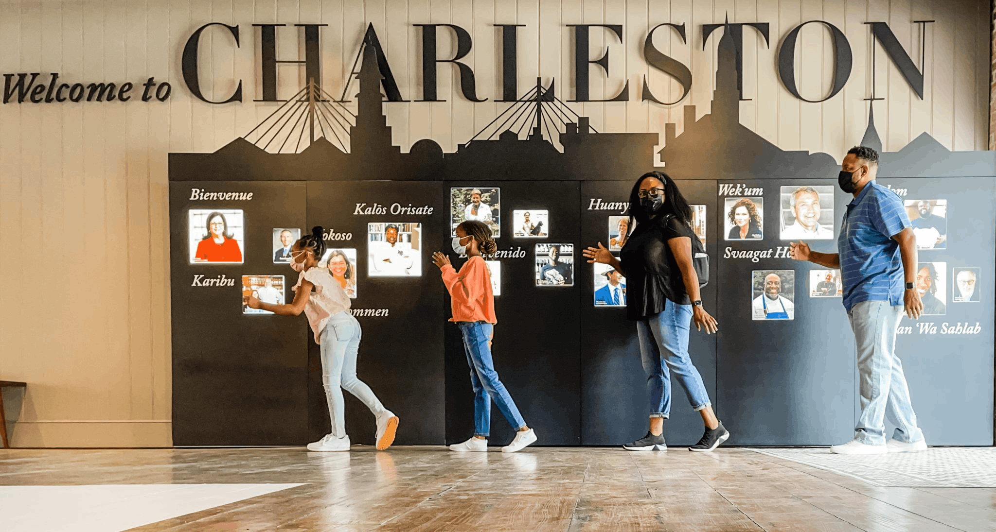 A family of four walking in a line in front of a "Welcome to Charleston" display with a city skyline silhouette and multi-language greetings.