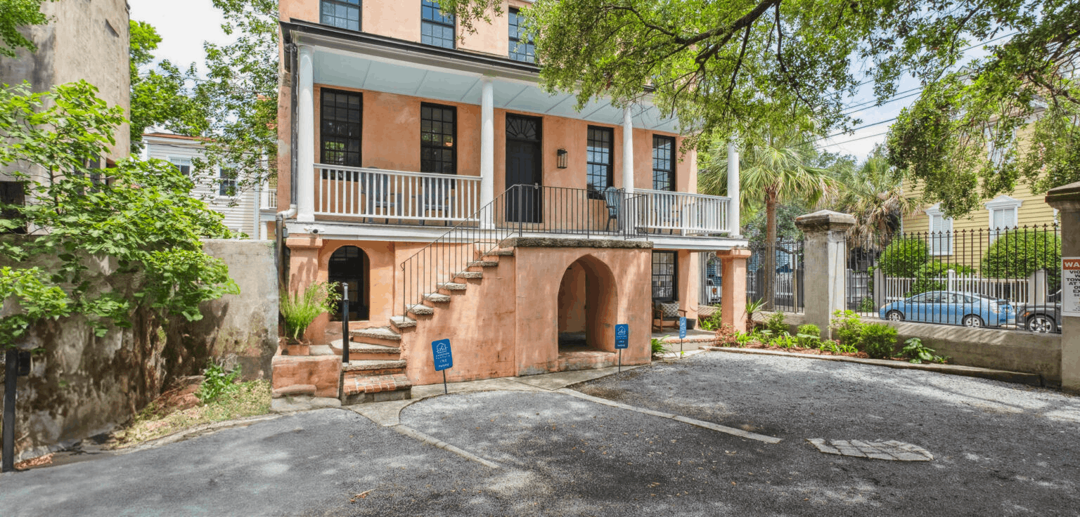 Exterior view of 179 St. Philip E, a historical three-story southern residence in downtown Charleston with a peach stucco facade, white balconies, and outdoor parking area.