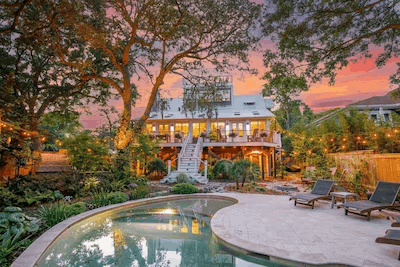 Luxury multi-story beach house with an illuminated balcony, private curved swimming pool, and lounge chairs surrounded by lush green trees under a vibrant sunset sky.