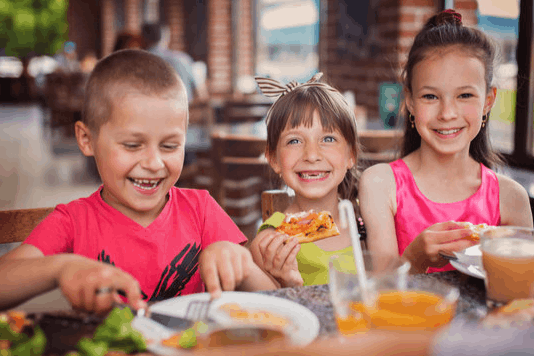 Three smiling children sitting at a restaurant table eating pizza and salad while drinking orange juice.