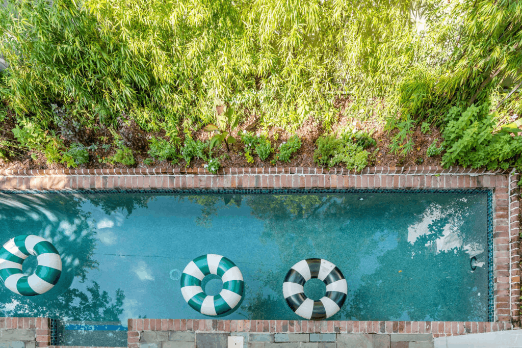 Top-down view of a narrow swimming pool with clear blue water, brick borders, three striped inflatable swim rings floating inside, and a lush green bamboo hedge in the background.